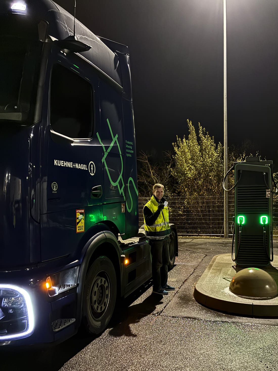 Batterieelektrischer Lkw nachts an der Schnellladesäule am IDS-Zentral-Hub, davor ein Mitarbeiter in Warnweste